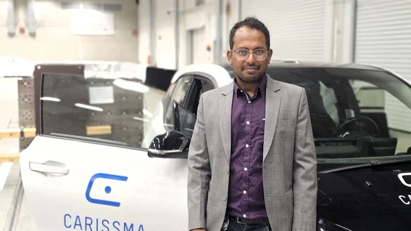 For more vehicle safety: Gerald Joy Sequeiras uses the CARISSMA hall for his driving tests (Photo: THI). Portrait mit Mann im Vordergrund, hinter Ihm ein Auto mit "CARISSMA"-Label auf der offenen Beifahrertüre