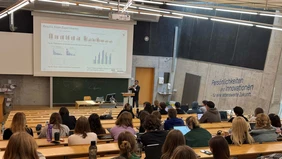 View from the back of the lecture hall, a presentation on the screen at the front, a speaker in a suit at the lectern, the students can be seen from behind