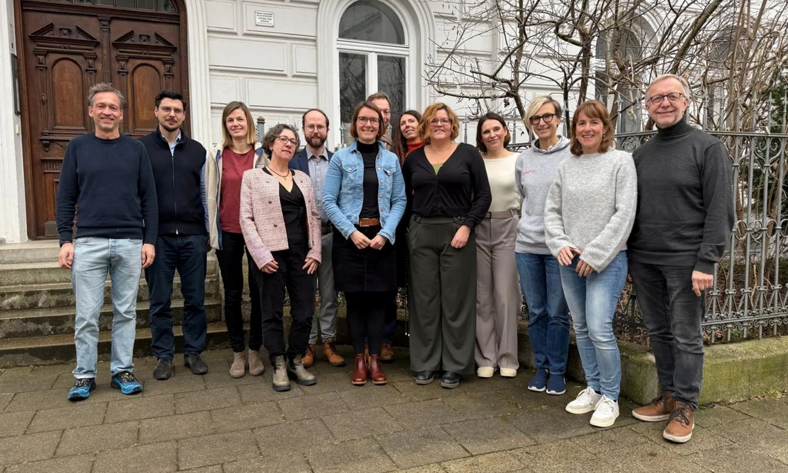 13 Personen stehen für ein Gruppenfoto vor einem Haus