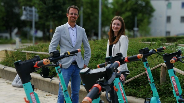 Ein Mann und eine Frau stehen hinter 5 türkisen E-Rollern. 