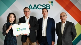 Four people are smiling into the camera, two of them are holding up a sign that reads: “AACSB accredited”.