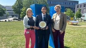 : drei Personen stehen auf einer Wiese und halten eine Plakette ins Bild 